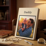 A hardcover photo book standing upright on a wooden desk, printed with the name 'Daddy' at the top and featuring a custom photo of a man holding a baby. The book is styled in a warm, cozy home office setting with a lamp, glasses, and a notebook nearby.